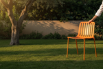 An orange slatted outdoor chair on a green lawn, with a person's arm and hand resting on the backrest near a tree and beige wall in the background.